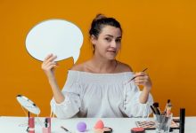 pleased-beautiful-girl-sits-table-with-makeup-tools-holds-chat-bubble-makeup-brush-isolated-orange-wall_141793-68495-3484821