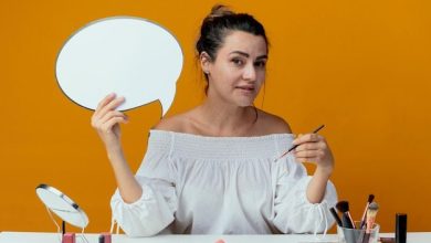 pleased-beautiful-girl-sits-table-with-makeup-tools-holds-chat-bubble-makeup-brush-isolated-orange-wall_141793-68495-3484821